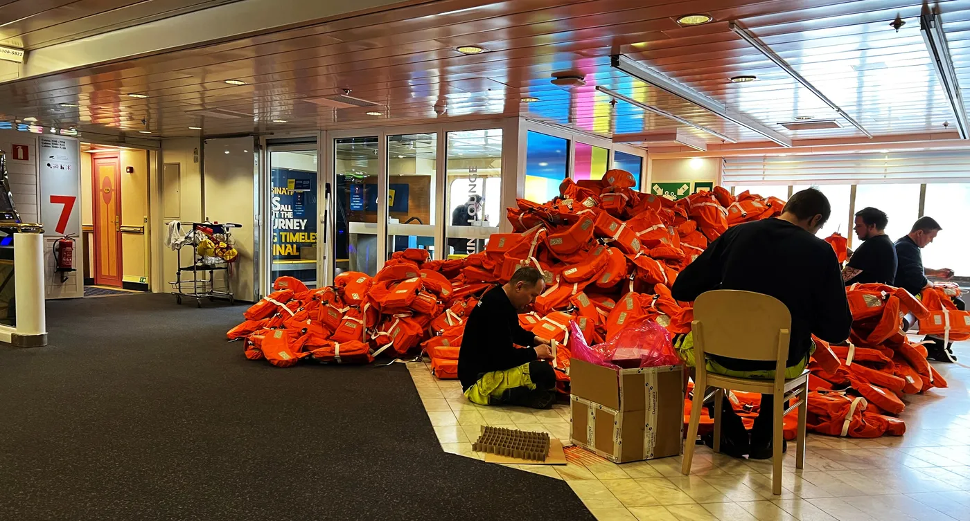 Tripulação revisa coletes salva-vidas laranja no salão de um navio de passageiros